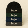 Stack of four embroidered beanies in black, bottle green, deep royal, and charcoal, displayed on a neutral background